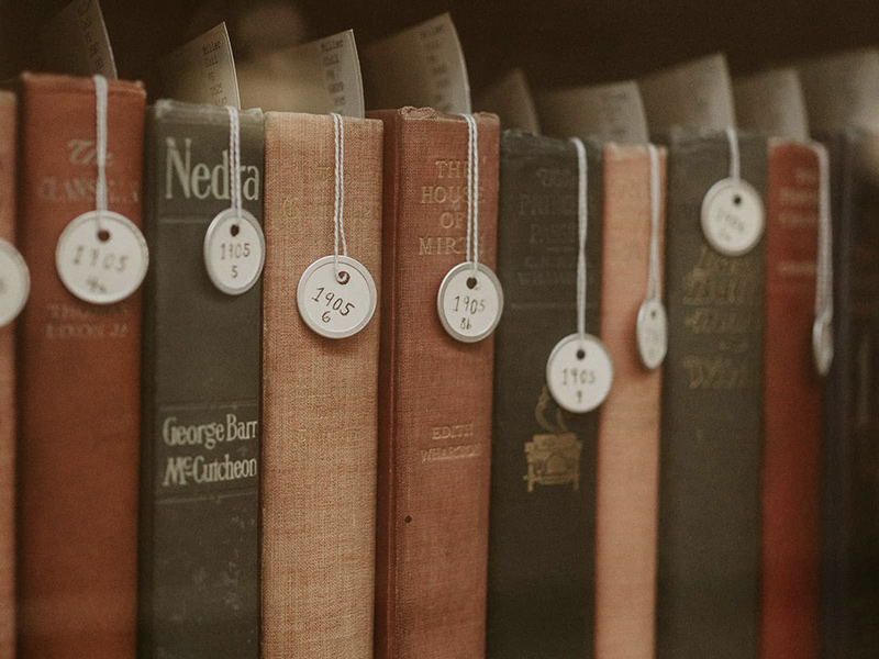 Nine books lined up on a shelf, each with circular tags labeled with the the number “1905”